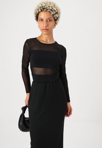 Juoda ilgomis rankovėmis suknelė su permatomu viršutiniu sluoksniu ir tvirtu sijonu. Lygus paviršius, prigludusi juosmens dalis ir raištelio detalė. Su derančia juoda pinigine.