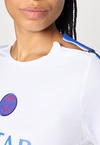 Haut de sport blanc avec des accents bleus, présentant une fermeture éclair à l'épaule et un logo circulaire avec le texte "Paris". Texture lisse.