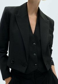 Sort cropped blazer med peak-lapels, to knapper og strukturerede skuldre. Den matchede vest nedenunder har tilsvarende knapper og tekstur.