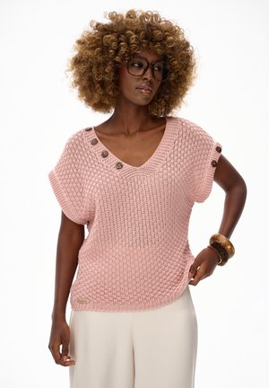 Kobieta o kręconych włosach i okularach, ubrana w różowy dzianinowy sweter na guziki oraz beżowe spodnie, pozująca z ręką na biodrze i drewnianymi bransoletkami.