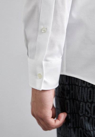 White long-sleeve shirt with a textured finish, featuring two buttons on the cuff. Worn with patterned dark fabric below.