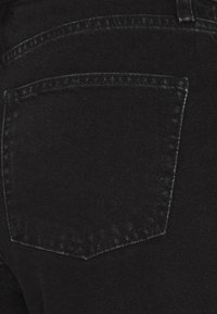 Schwarzer Denim-Stoff mit einer sichtbaren Gesäßtasche, die an der Oberkante spitz zuläuft und dezente Nähdetails aufweist. Die Textur wirkt glatt und robust.