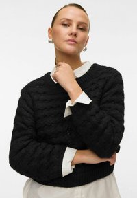 Svart stickad tröja med en strukturerad vågmönster, rund halsringning och ribbade muddar, bärs över en vit skjorta med krage.