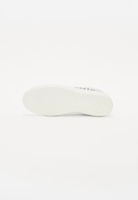 Bílá podrážka tenisky s trojúhelníkovým texturovaným vzorem na čistě bílém pozadí, umístěná na boku.