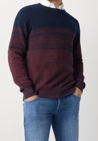 Hombre vistiendo un suéter de punto ombré en azul marino y burdeos sobre una camisa blanca con cuello, con las manos en los bolsillos de unos jeans azules, contra un fondo blanco.