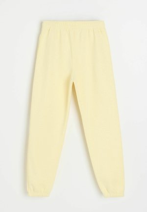 Jasnożółte elastyczne spodnie dresowe z gumką w pasie i przy kostkach, ułożone na płasko, widok z przodu z marszczonymi mankietami i gładką fakturą.