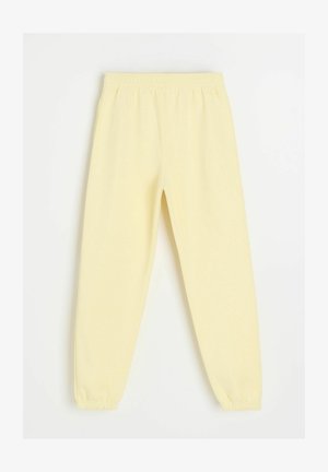 Jasnożółte elastyczne spodnie dresowe z gumką w pasie i przy kostkach, ułożone na płasko, widok z przodu z marszczonymi mankietami i gładką fakturą.