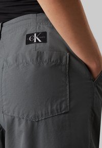 Sive hlače s stražnjim džepom na kojem je crni logo patch Calvin Klein Jeans. Tkanina je glatka s mat površinom i urednim šavovima.