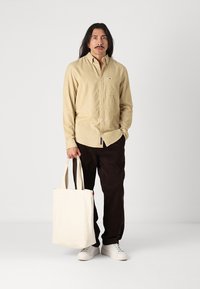 Beige kordflortskjorta med knappar och bröstficka, bruna kordbyxor och vita sneakers. Håller en krämfärgad toteväska med röd accent.
