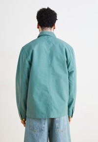 Veste oversized teal avec fermeture à bouton-pression, en tissu légèrement texturé, et un détail de couture au dos, associée à un jean en denim bleu clair.
