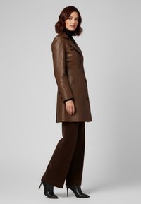 Bruine leren jas met een getailleerde silhouet, dubbele knopen en zijzakken, gecombineerd met donkerbruine wijde broek en zwarte hakken.