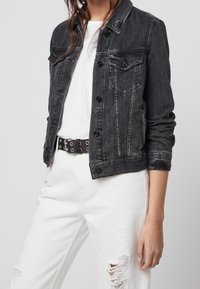 Černá džínsová bunda s knoflíkovým zapínáním, dvěma náprsními kapsami a ohrnutými rukávy. Nositá přes bílý tričko a kombinovaná s obnošenými bílými džíny.