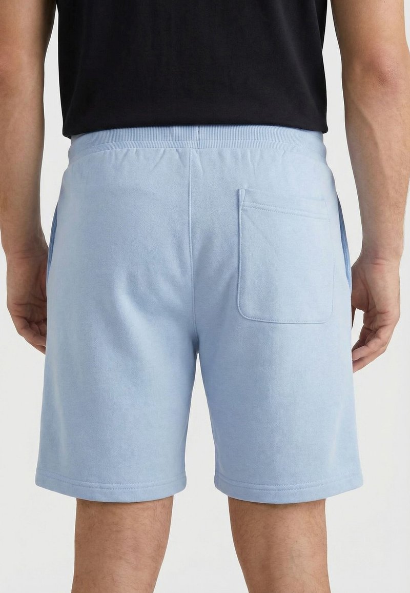 Homme portant un short bleu clair avec une taille élastique et une seule poche arrière, vu de dos sur un fond uni.