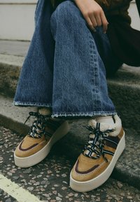 Plattform-Sneaker mit einem Obermaterial aus beigem Wildleder, dunklen Akzenten, Schnürverschluss, Fellfutter und einer weißen strukturierten Sohle, kombiniert mit blauen Schlagjeans.