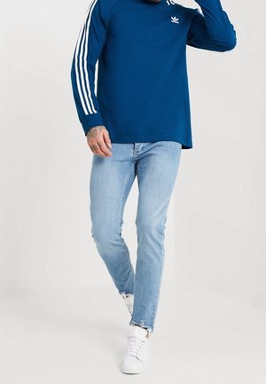 Niebieska koszula z długim rękawem z białym wzorem z trzema paskami na rękawach, w połączeniu z jasnoniebieskimi obcisłymi dżinsami i białymi trampkami.