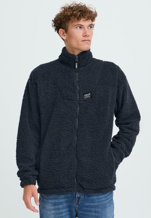 Schwarze Fleecejacke mit hohem Kragen, Reißverschluss vorne, strukturiertem Außenmaterial und einem kleinen Markenetikett auf der Brust. Getragen über blauen Jeans.