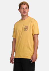 Geel katoenen t-shirt met korte mouwen, ronde halslijn en een cirkelvormig rood grafisch logo op de borst. Eenvoudige, ontspannen pasvorm.