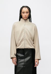 Μπεζ ζακέτα με φερμουάρ, ψηλό γιακά και φαρδιά μανίκια, συνδυασμένη με μαύρη δερμάτινη φούστα με λείο υφή.