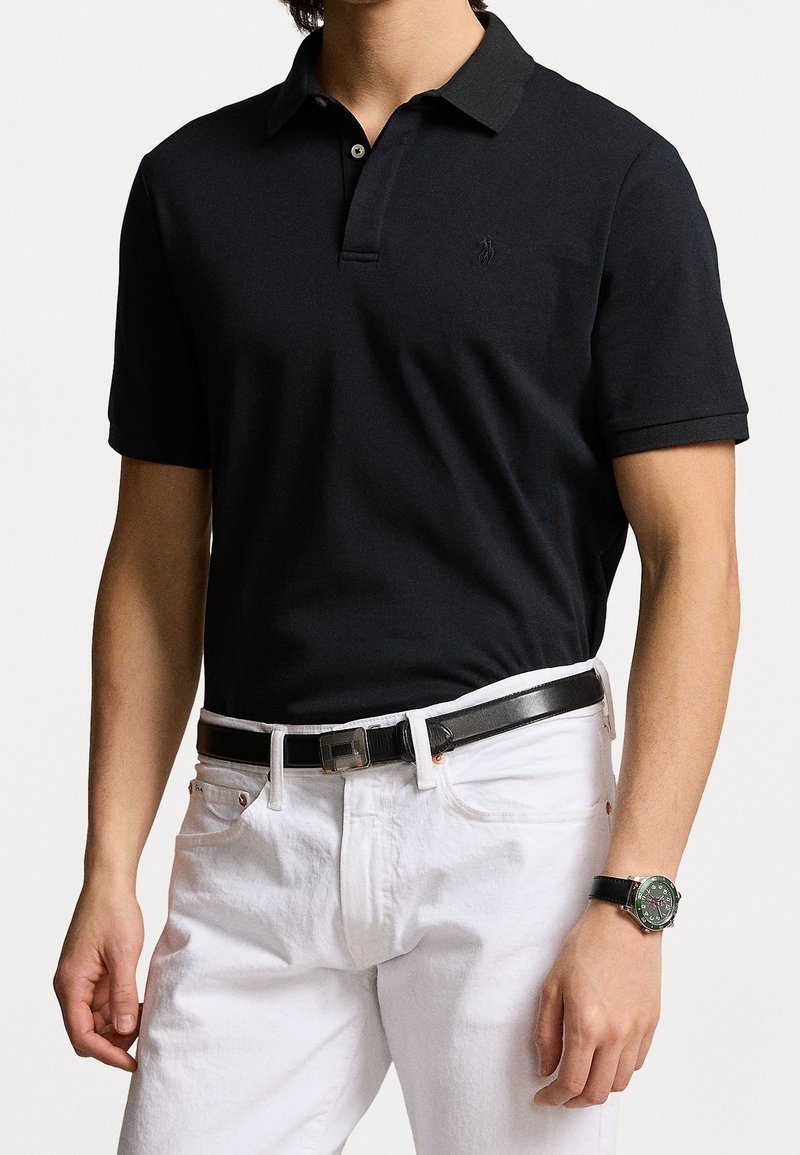 Homme portant un polo noir à manches courtes, un pantalon blanc, une ceinture noire et une montre au poignet sur un fond clair uni.