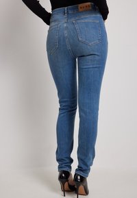 Vaqueros ajustados de mezclilla azul claro con cintura alta, cinco bolsillos y una etiqueta de marca de cuero marrón. Combinados con zapatos de tacón alto negros.