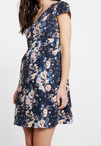 Femme portant une robe à manches courtes florale bleu marine avec un décolleté en V et une jupe évasée.