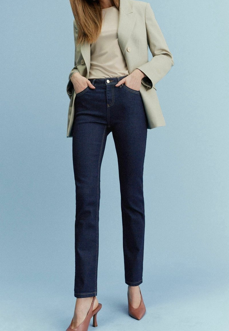 Mørkeblå jeans med slim fit, parret med en tan-farvet top og en lys grå blazer. Brune højhælede sko fuldender outfittet.