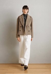 Veste à double boutonnage en laine marron portée sur un col roulé charcoal, associée à un pantalon large blanc et des chaussures marron, debout sur un sol en bois.