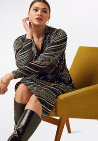 Långärmad klänning i rutig mönster med blå, bruna och krämfärgade nyanser; i kombination med höga svarta läderstövlar. Gul accentstol i bakgrunden.