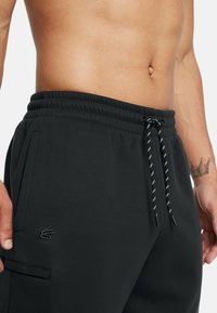 Črne trenirke z elastičnim pasom, podrobnostjo z vrvico, stranskimi žepi in diskretno izvezenim logotipom na stegnu. Gladka tekstura tkanine.