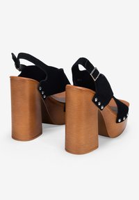 Sandalias de plataforma de ante negro con un tacón grueso de madera marrón, con tachuelas plateadas y una correa ajustable en el tobillo.