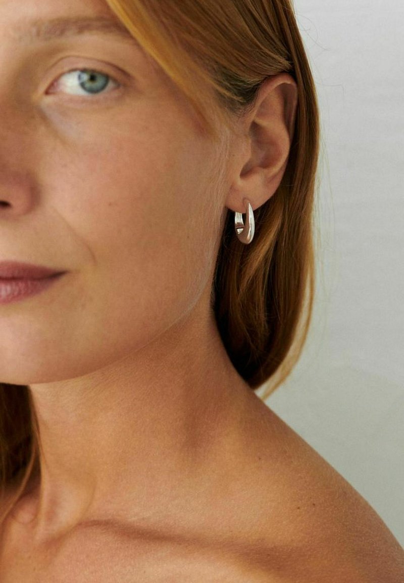 Moterys su šviesia oda ir žalia akimi, dėvinčia mažą sidabrinį lankinį auskarą, matoma nuo skruosto iki peties ant paprasto fono.