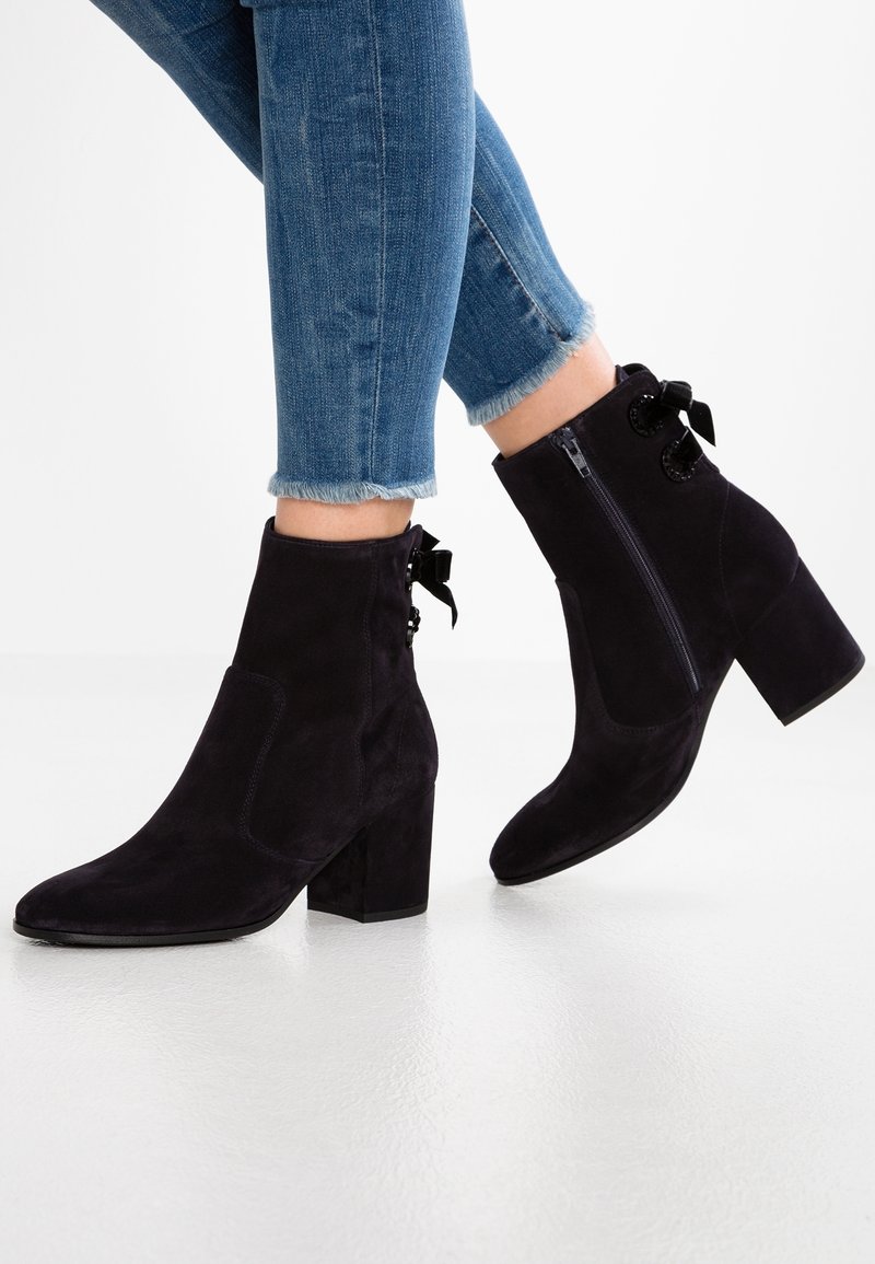 Personne portant un jean skinny bleu et des bottines en daim noir à talons carrés avec des nœuds décoratifs sur les côtés.