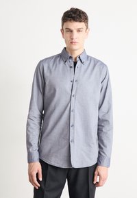 Button-up shirt in light blue chambray fabric; features a pointed collar, long sleeves, and subtle embroidered logo on the chest.