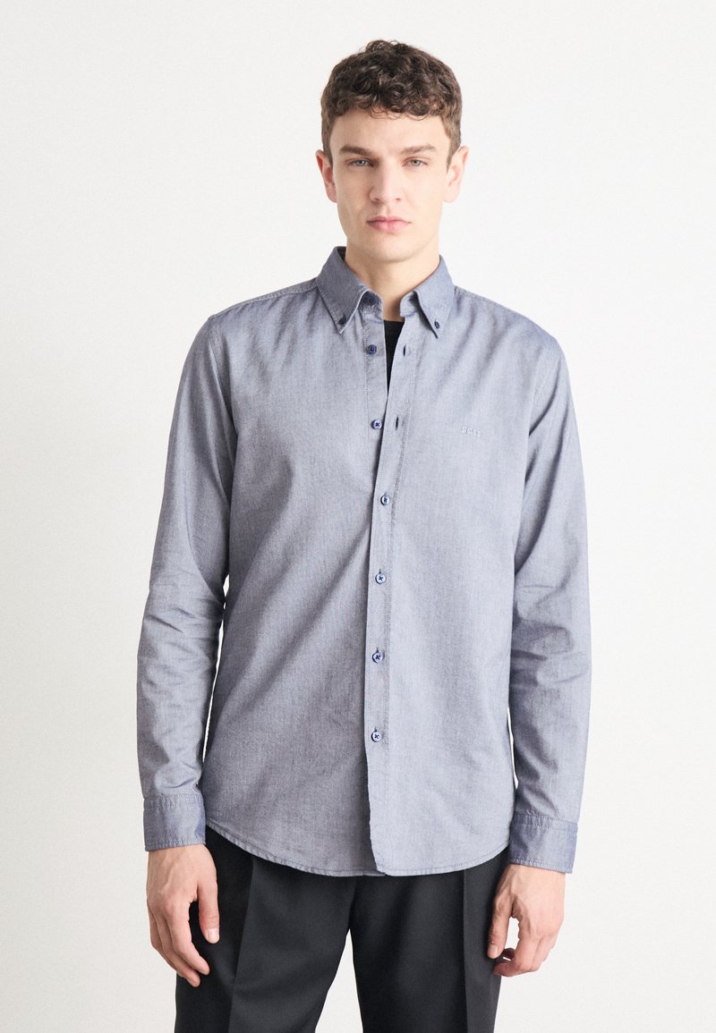 Button-up shirt in light blue chambray fabric; features a pointed collar, long sleeves, and subtle embroidered logo on the chest.