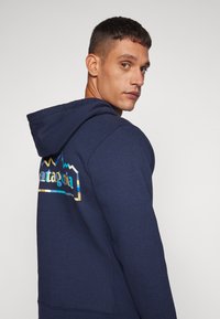 Hanorac bleu marin cu un finisaj texturat, având un logo multicolor cu munte pe spate, buzunar frontal tip kangaroo și glugă ajustabilă.
