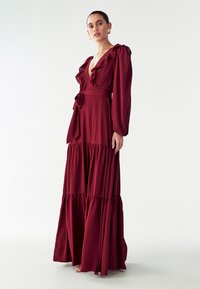 Ilga burgundi spalvos suknelė su volanais, gilia V formos iškirpte, ilgomis rankovėmis ir prigludusia juosmens dalimi. Platėjanti, pakopinė sijonas su tekanti medžiaga ir šonine plyšiu.