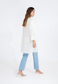 Top lungo bianco e strutturato con maniche a tre quarti, abbinato a jeans a gamba dritta blu chiaro e sandali con tacco alto bianchi e con cinturino.