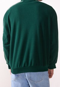 Giacca in fleece verde con colletto alto e strisce bianche sui lati, dal taglio rilassato e con vita elastica. Design semplice e sobrio.