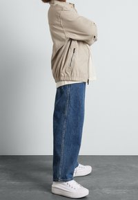 Beżowa kurtka zapinana na zamek z elastycznymi mankietami, zestawiona z niebieskimi dżinsami z wysokim stanem i białymi trampkami. Prosty, luźny krój.