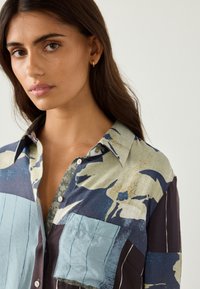 Chemise boutonnée présentant un motif multicolore avec des nuances de bleu, vert et beige. Tissu texturé avec une grande poche.
