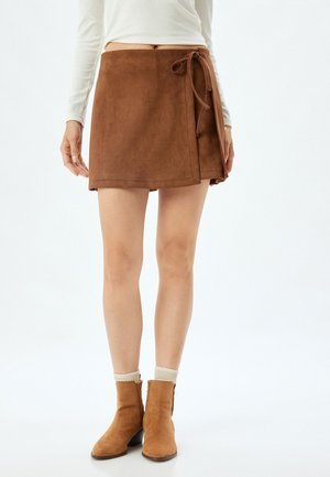Jupe enveloppante en faux suède marron avec un nœud sur le côté, présentant un design épuré et une surface lisse. Jambes visibles, associée à des bottines.