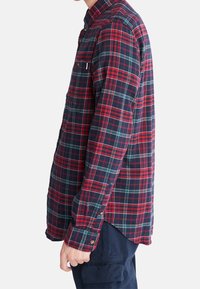 Camicia a maniche lunghe a quadretti rossi e blu con colletto abbottonato, taschino sul petto singolo e orlo arrotondato; presenta linee verdi a contrasto.