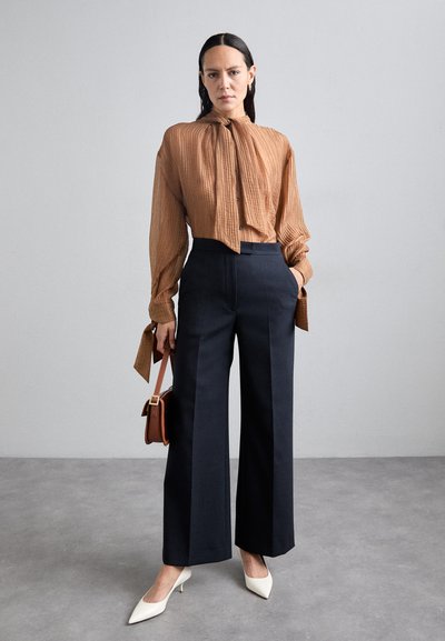 Brun rillede bluse med båndhals, parret med marineblå vide bukser. Modellen bærer en lille brun håndtaske og har hvide spidse hæle på.