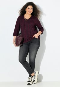 Femme souriante avec des cheveux bouclés, portant un haut à motifs sombres, un jean noir, des baskets robustes, des bijoux en or, et tenant un sac texturé foncé.