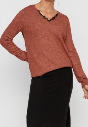 Femme portant un pull col en V de couleur rouille avec une bordure en dentelle noire et une jupe noire jusqu'aux genoux, sur un fond clair uni.