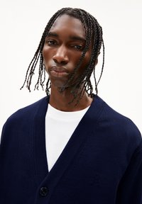 Cardigan bleu marine avec un col en V, présentant une fermeture à un seul bouton et des détails en tricot subtils, porté par-dessus un t-shirt blanc.