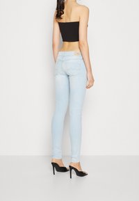 Jean skinny bleu clair avec un délavage subtil, présentant un détail de couture incurvé sur les poches arrière et une simple patch de logo beige à la taille.