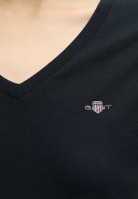 Gros plan d'un t-shirt noir à col en V avec un petit logo GANT brodé en blanc, rouge et bleu sur la poitrine.