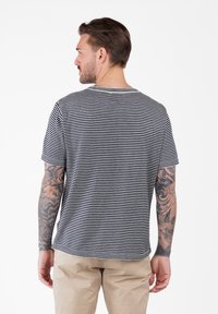 Hombre con cabello corto castaño que lleva una camiseta a rayas negras y blancas y pantalones beige, mostrando la espalda y brazos tatuados.