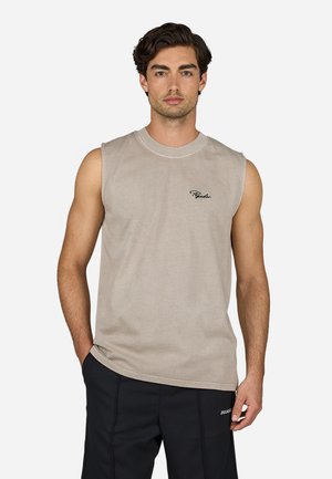 Sleeveless beige cotton tank top with round neckline and embroidered logo on left chest; model poses with hands in pockets, wearing dark shorts.